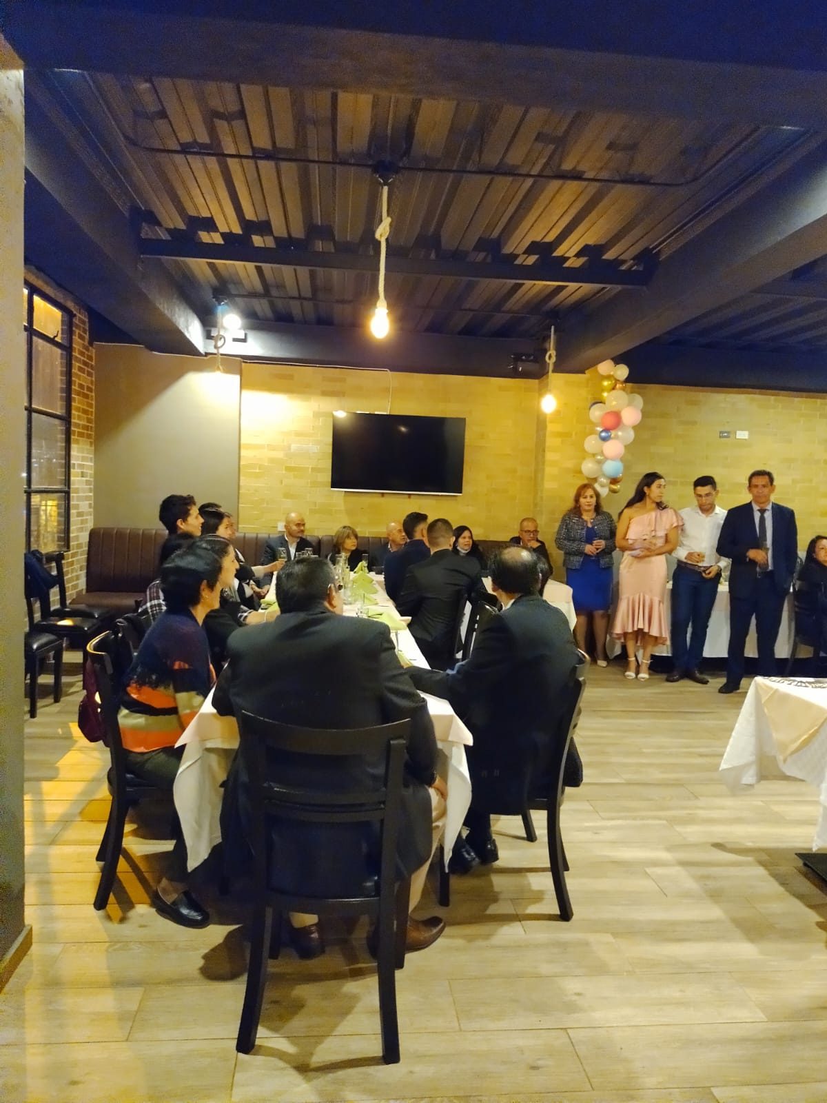 Invitados sentados a la mesa durante una celebración, vistos de espaldas, con globos al fondo