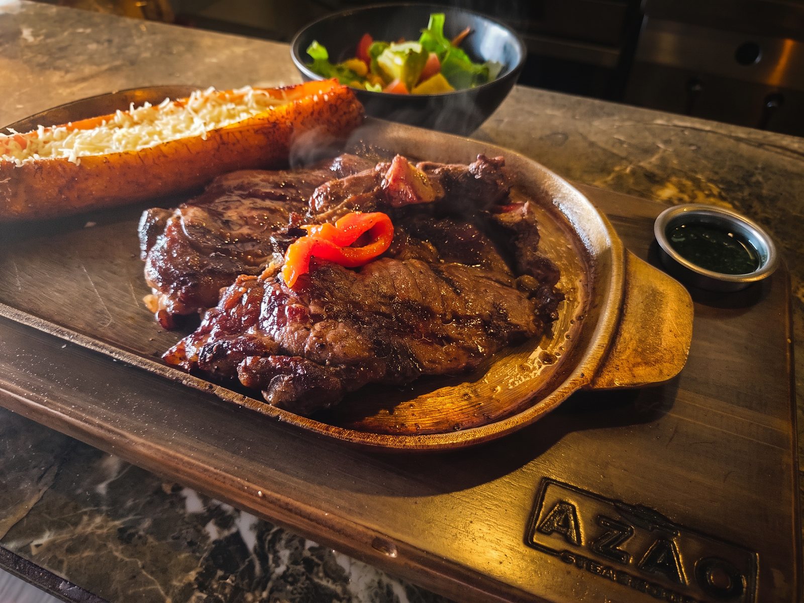 Bife a la parrilla con pimentón asado y maduro gratinado, servido en tabla de madera con el logo de Azao sobre el mesón, con la cocina al fondo