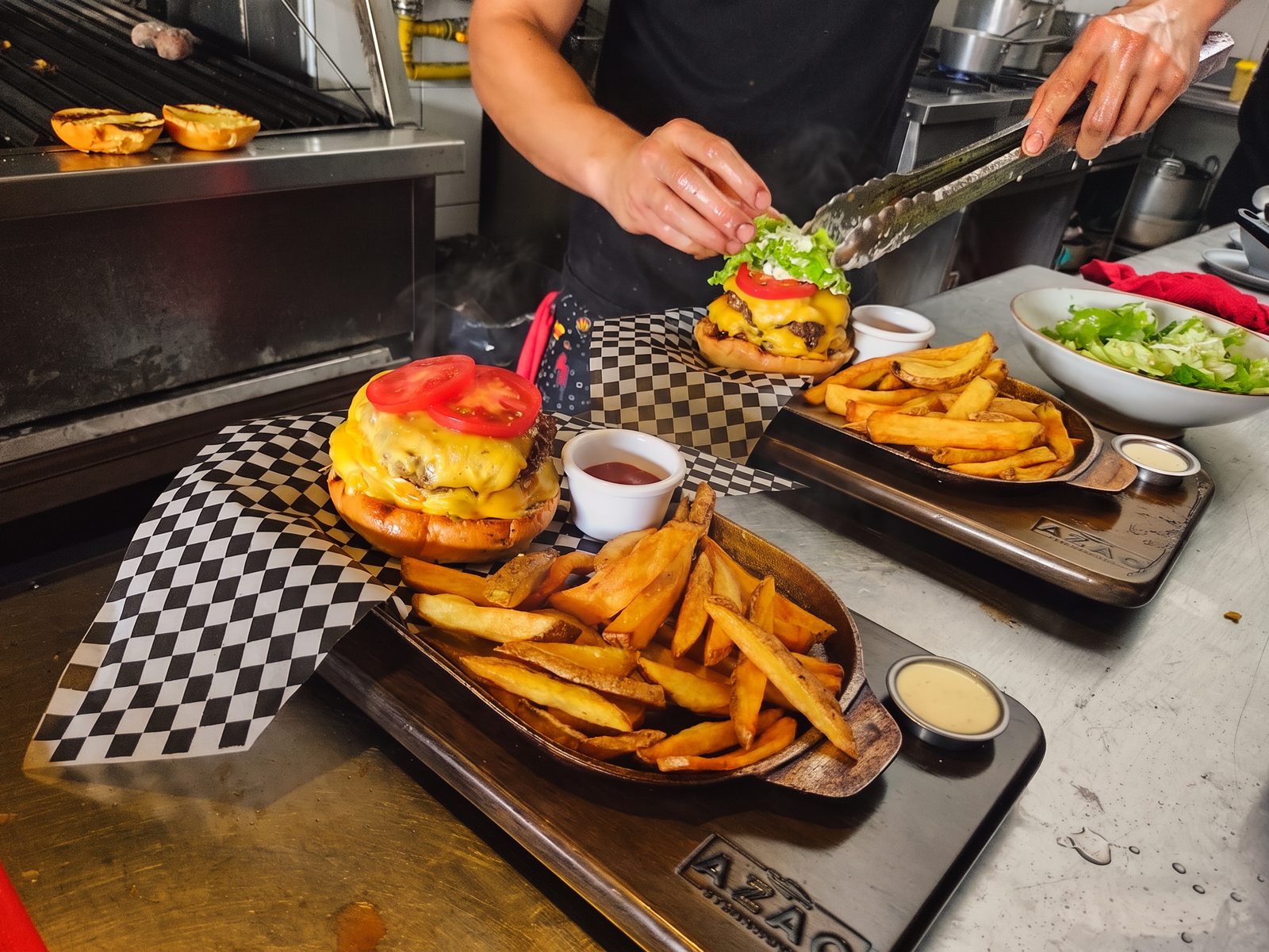 Cocinero arma una hamburguesa junto a la parrilla, se ven sus manos y el plato con papas