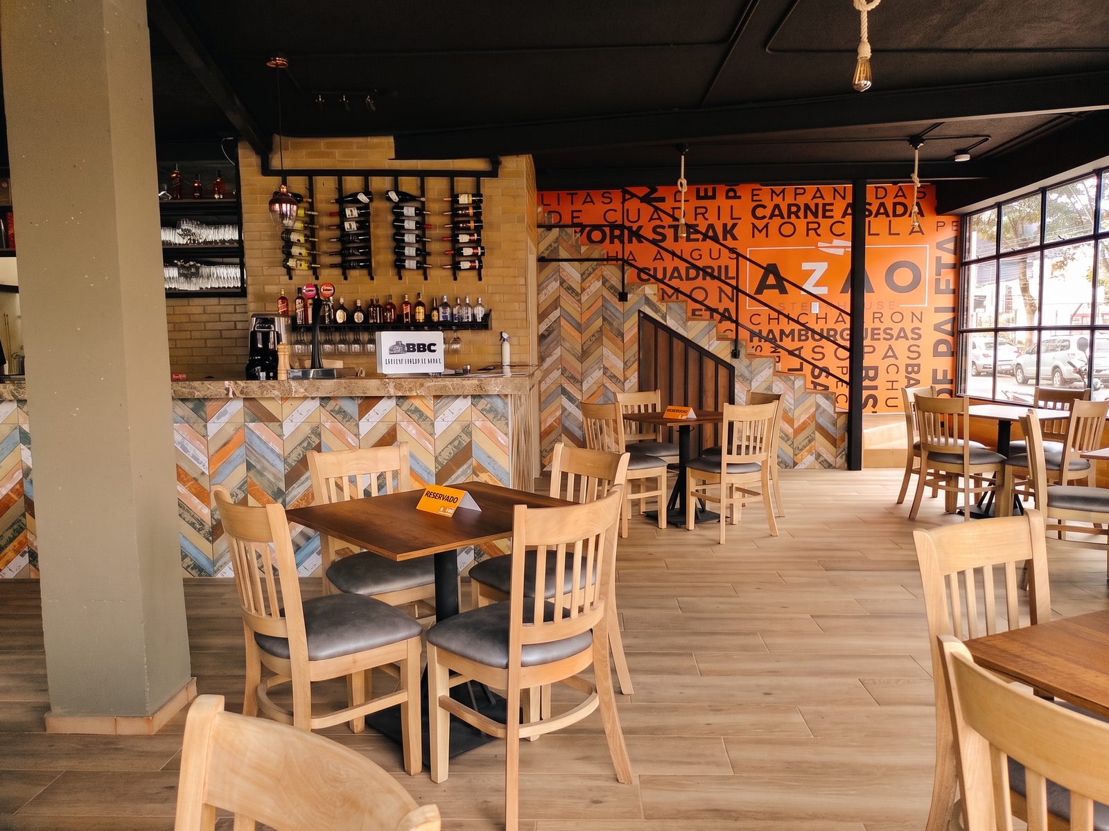 Salón con barra de baldosa en espiga, sillas de madera y pared naranja con nombres de cortes