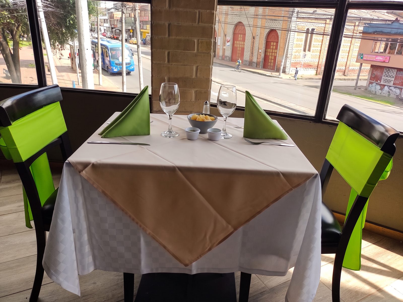 Mesa para dos junto al ventanal, con mantel, copas y moños verdes en las sillas